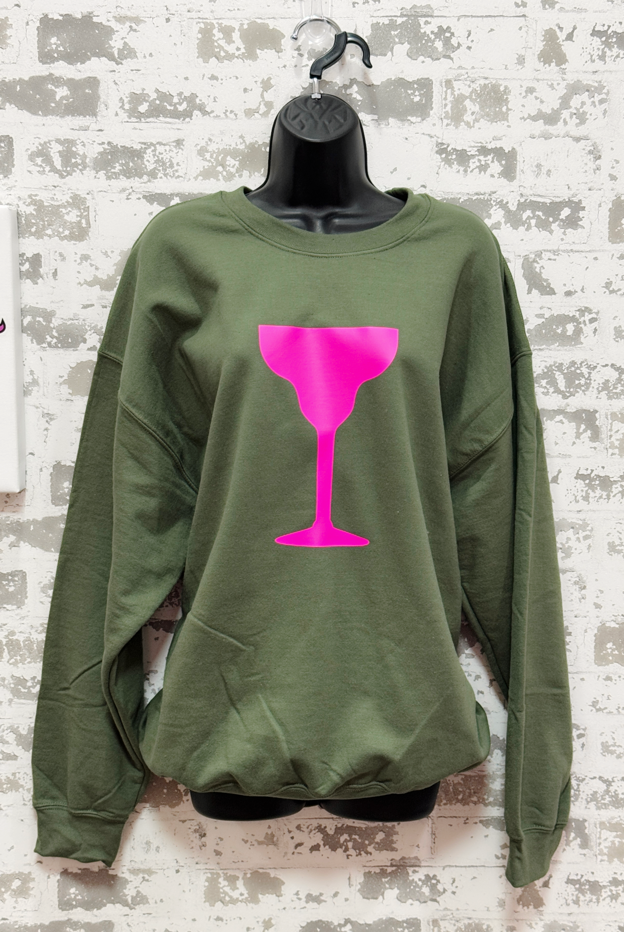 Green sweatshirt with a pink wine glass design on a mannequin against a textured wall.