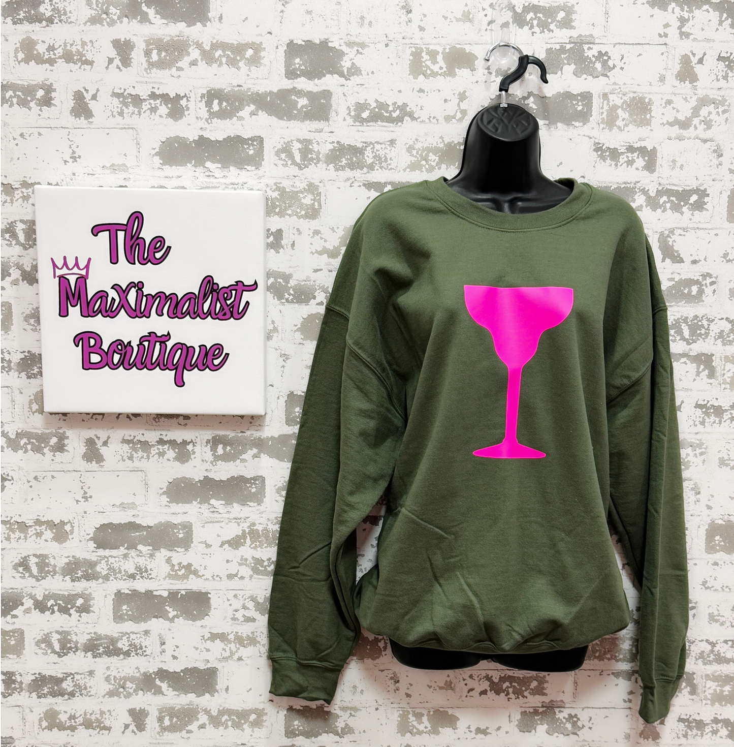 Green sweatshirt with pink wine glass design on a hanger against a textured wall with 'The Maximalist Boutique' sign.