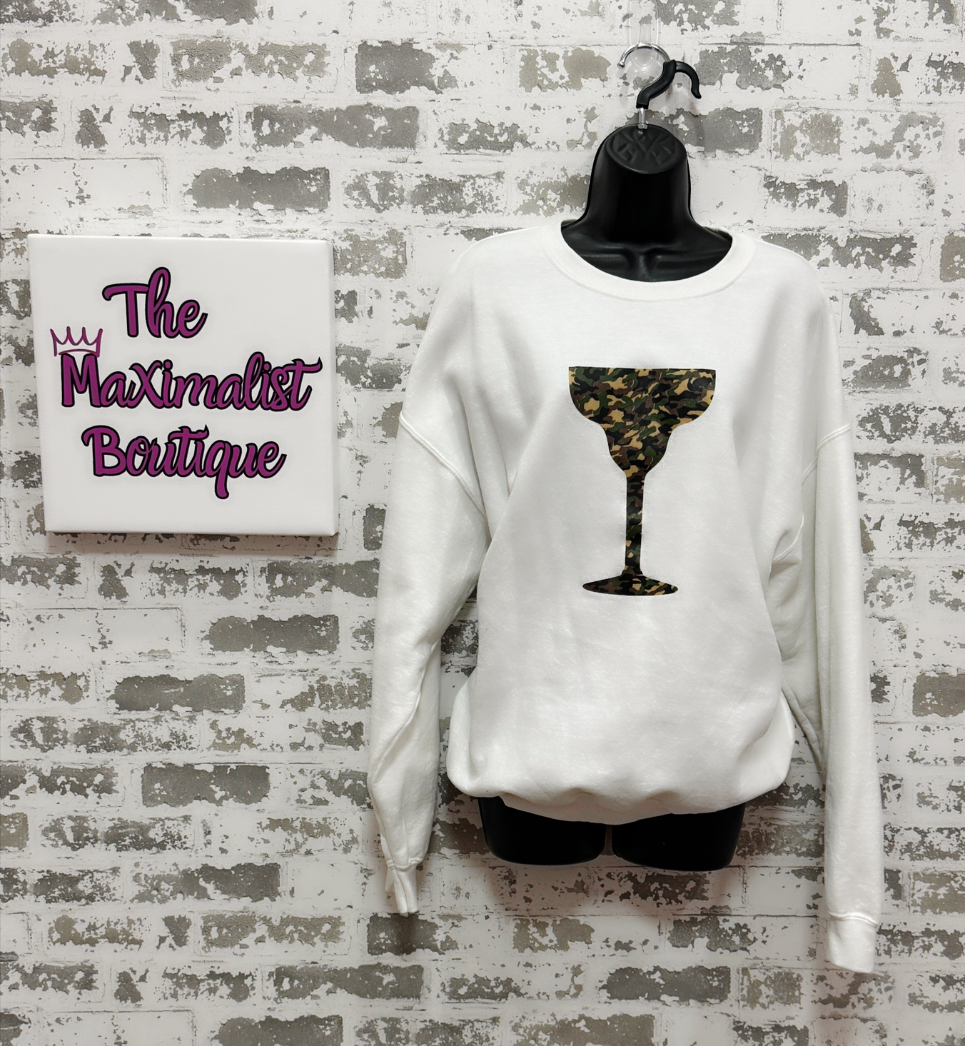 White sweatshirt with a camo design on a mannequin against a textured wall with 'The Maximalist Boutique' sign.