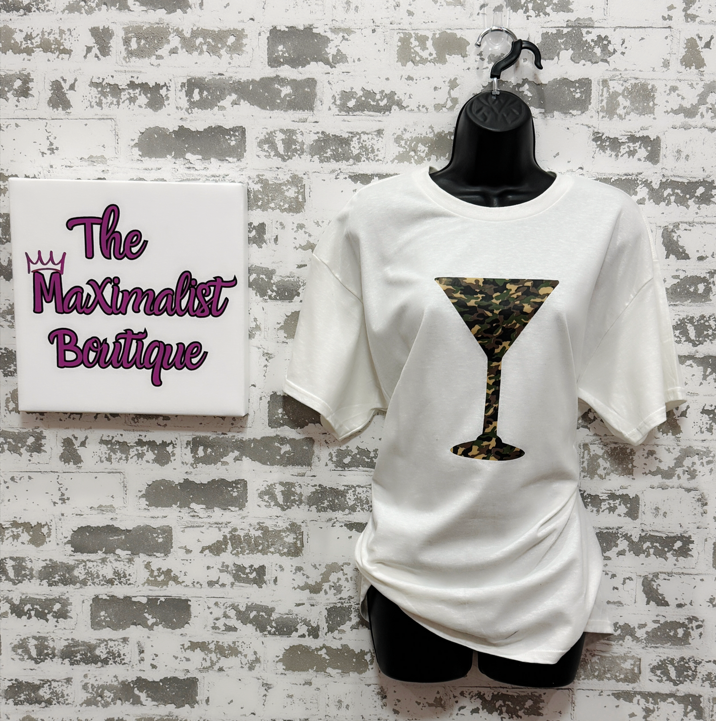 White t-shirt with camo print martini glass design on a mannequin against a brick wall with 'The Maximalist Boutique' sign.