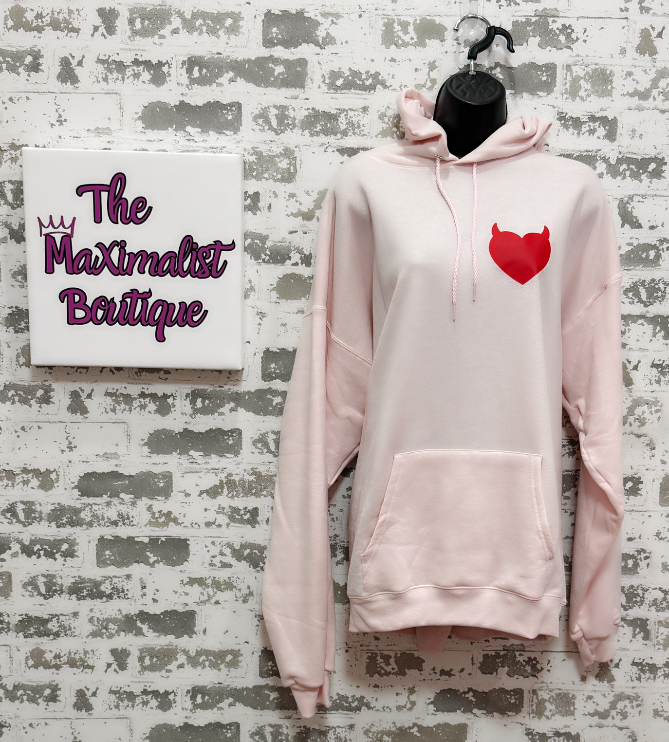 Pink hoodie with a red heart design on a textured wall with 'The Maximalist Boutique' sign.
