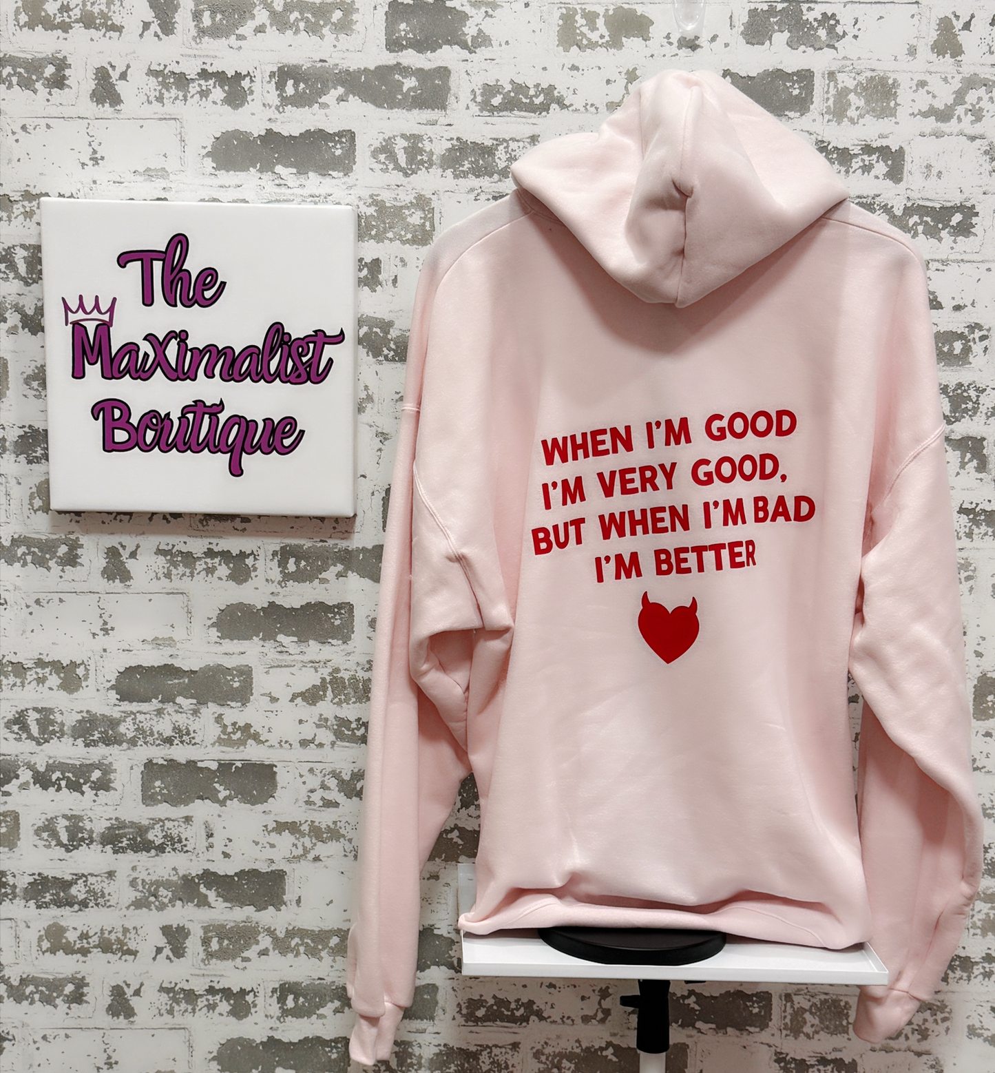 Pink hoodie with text on a textured wall, featuring 'The Maximalist Boutique'.