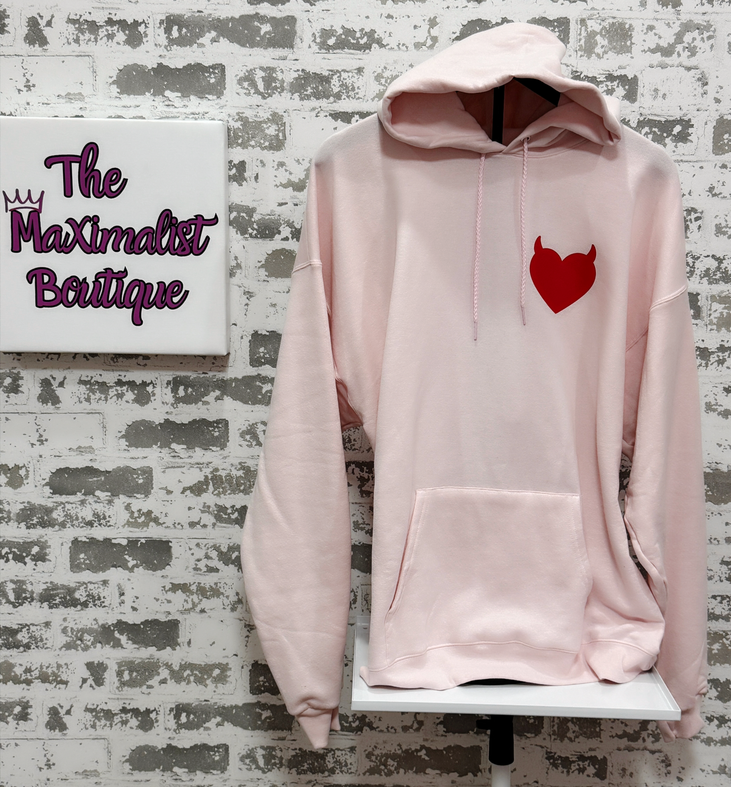 Pink hoodie with a red heart design on a brick wall background with 'The Maximalist Boutique' sign.