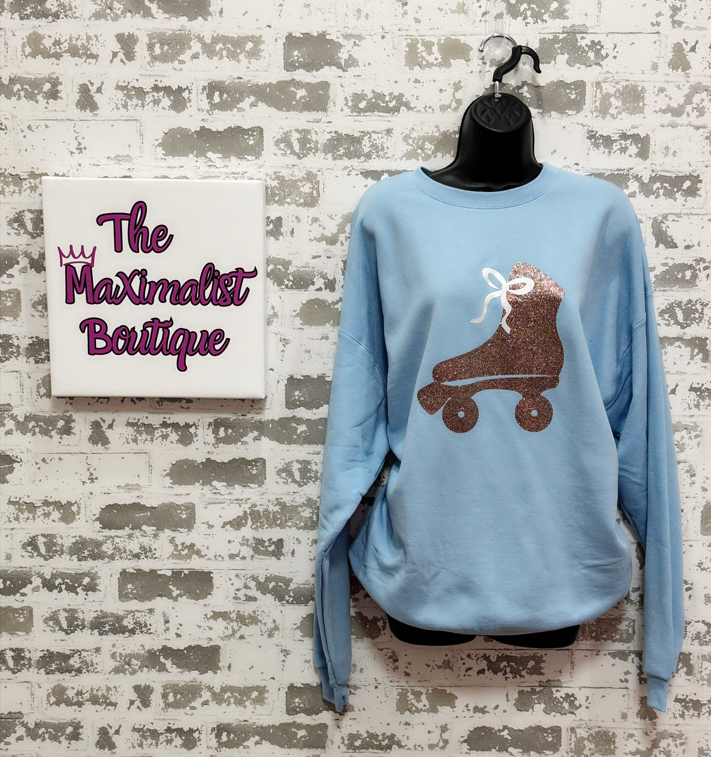 Light blue sweatshirt with a GLITTER roller skate design on a mannequin against a textured wall with 'The Maximalist Boutique' sign.