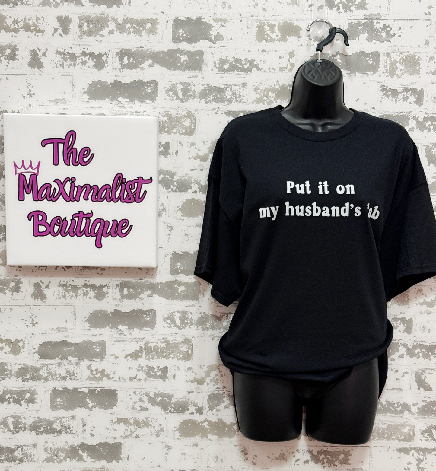 Black t-shirt with text on a mannequin against a textured wall with 'The Maximalist Boutique' sign.
