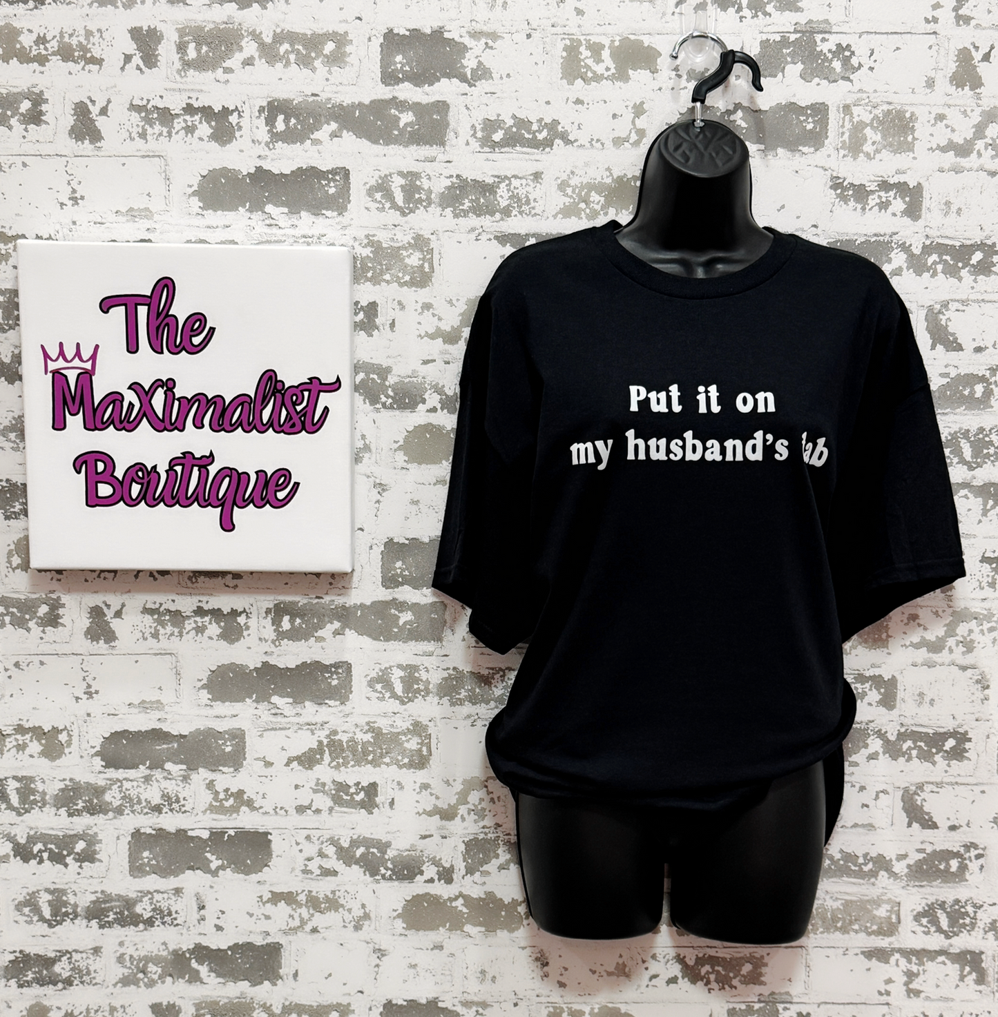 Black t-shirt with text on a hanger against a textured wall with 'The Maximalist Boutique' sign.