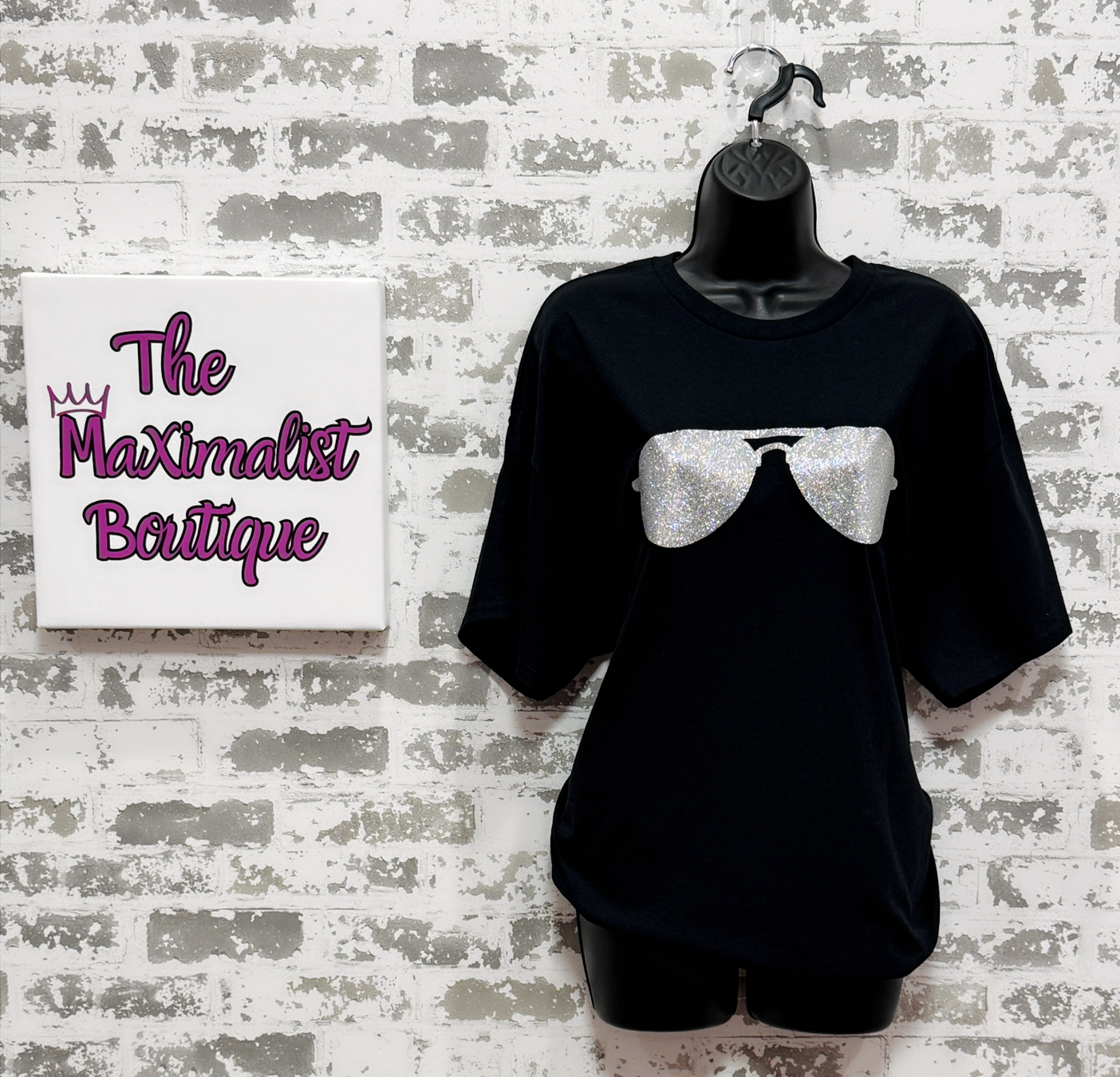 Black t-shirt with a white bow design on a mannequin against a textured wall with 'The Maximalist Boutique' sign.