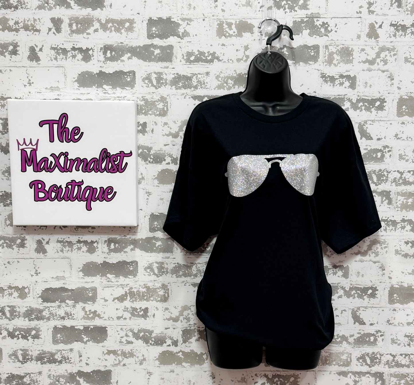 Black t-shirt with a graphic design on a mannequin against a textured wall with 'The Maximalist Boutique' sign.