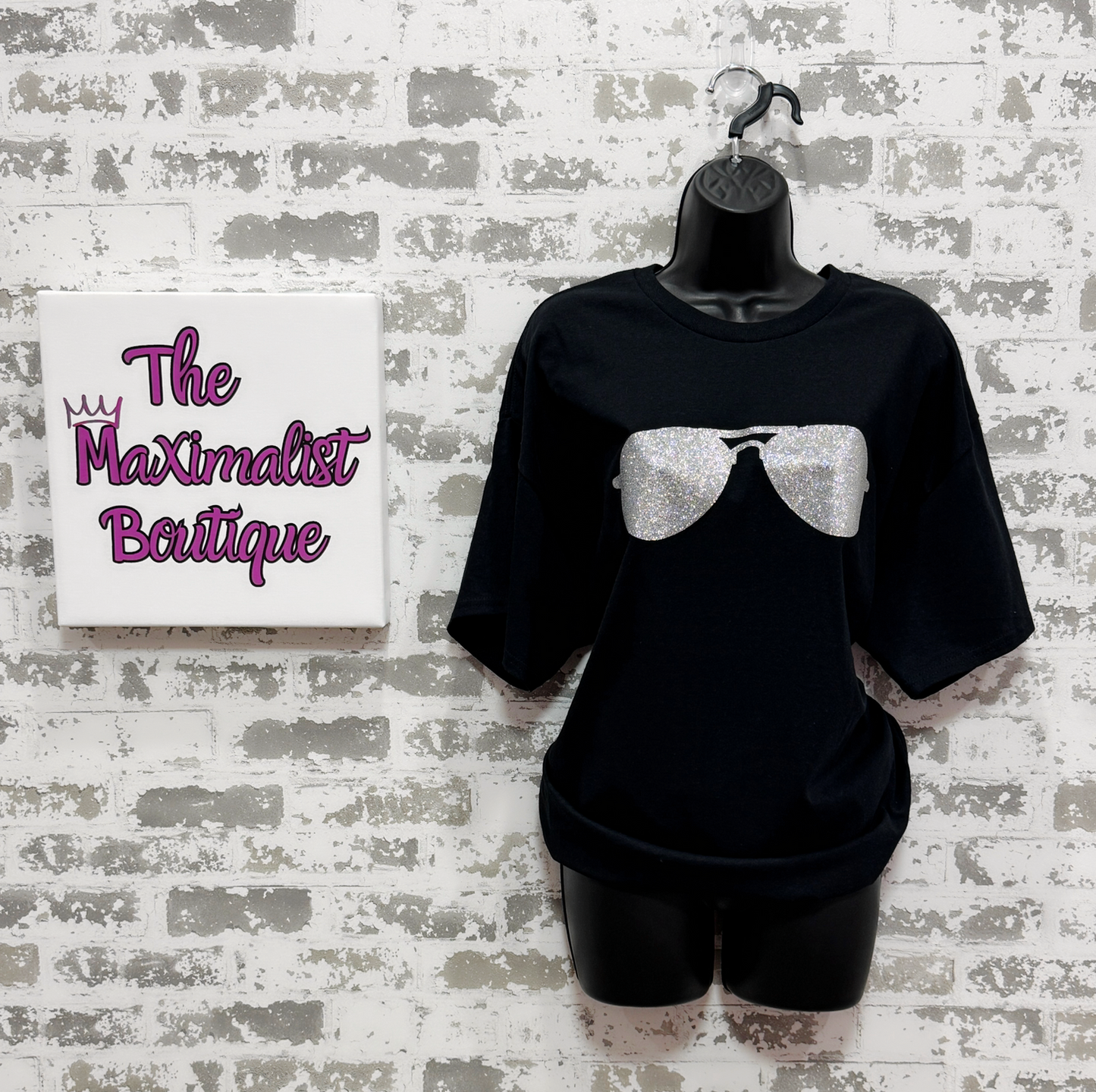 Black t-shirt with silver glitter bow design on a mannequin against a textured wall with 'The Maximalist Boutique' sign.