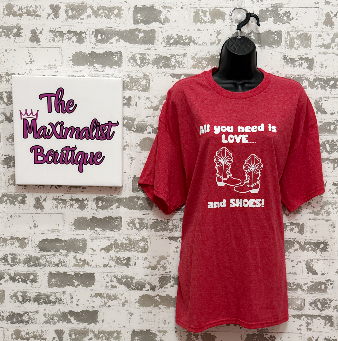 Red t-shirt with text 'All you need is love and shoes' on a hanger against a textured wall with 'The Maximalist Boutique' sign.