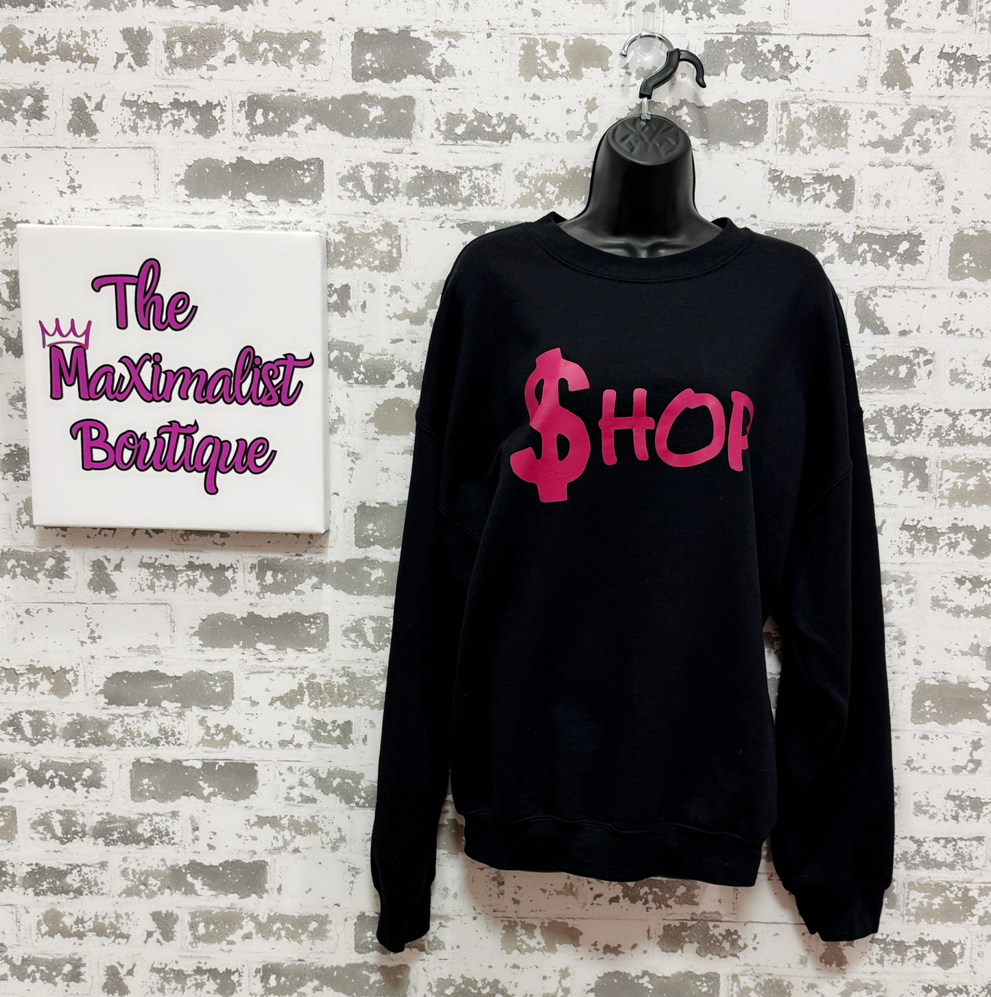Black sweatshirt with pink text 'SHOP' on a hanger against a textured wall with 'The Maximalist Boutique' sign.