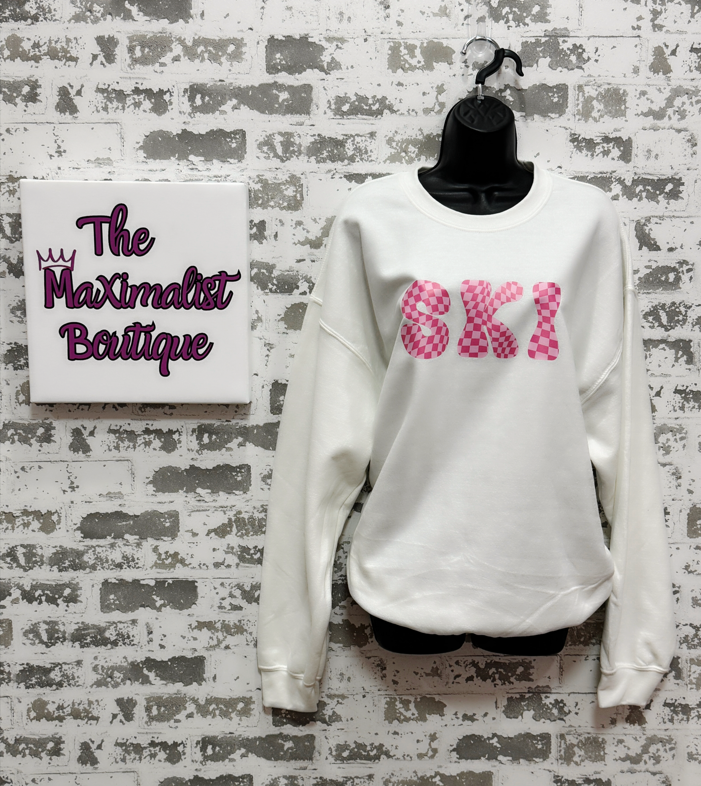 White sweatshirt with pink text on a mannequin against a textured wall with 'The Maximalist Boutique' sign.