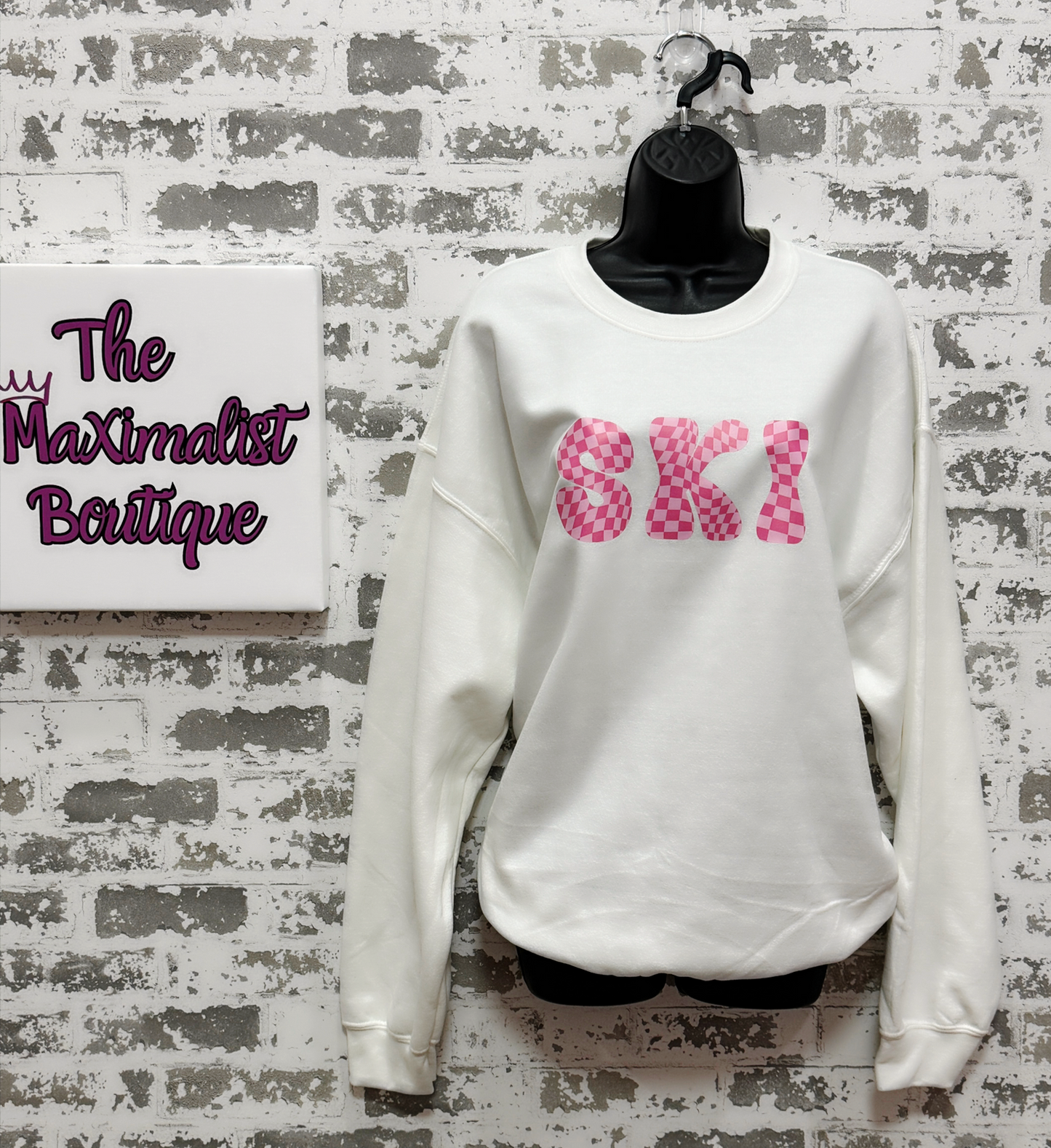 White sweatshirt with pink text on a textured wall with 'The Maximalist Boutique' sign.