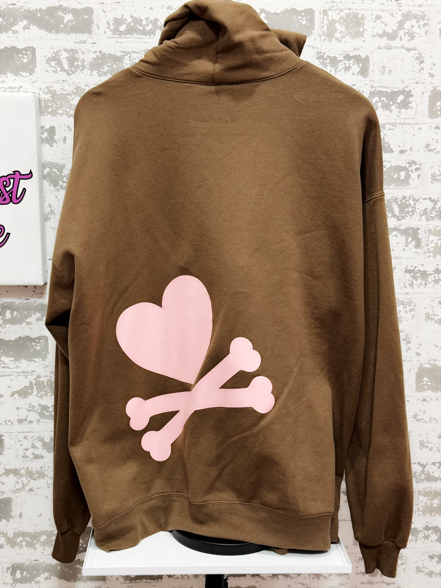 Brown hoodie with pink heart and bones design on a mannequin against a textured wall.