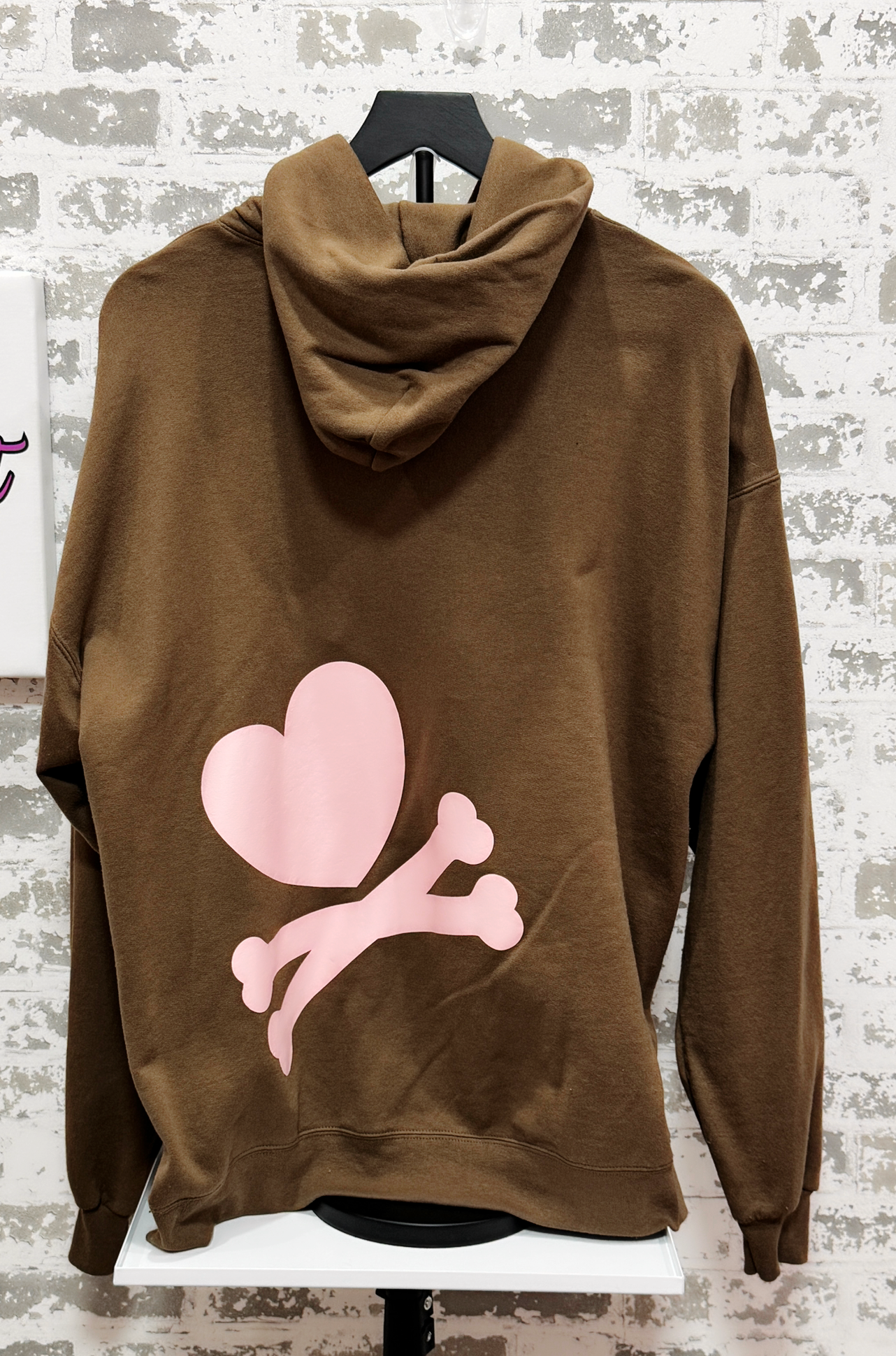 Brown hoodie with pink heart and bone design on a hanger against a textured wall.