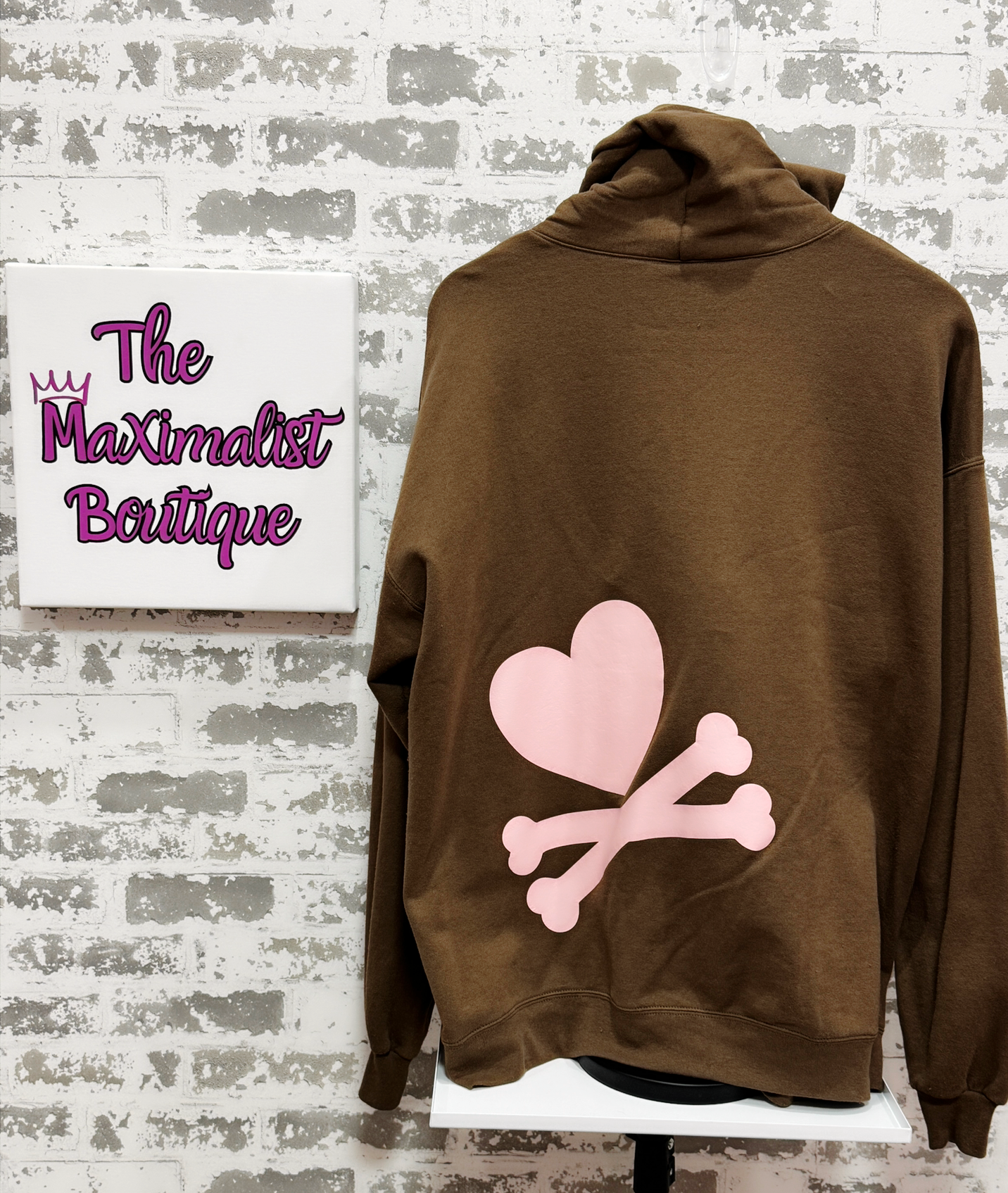 Brown hoodie with pink heart and bones design on a white stand against a textured wall with 'The Maximalist Boutique' sign.