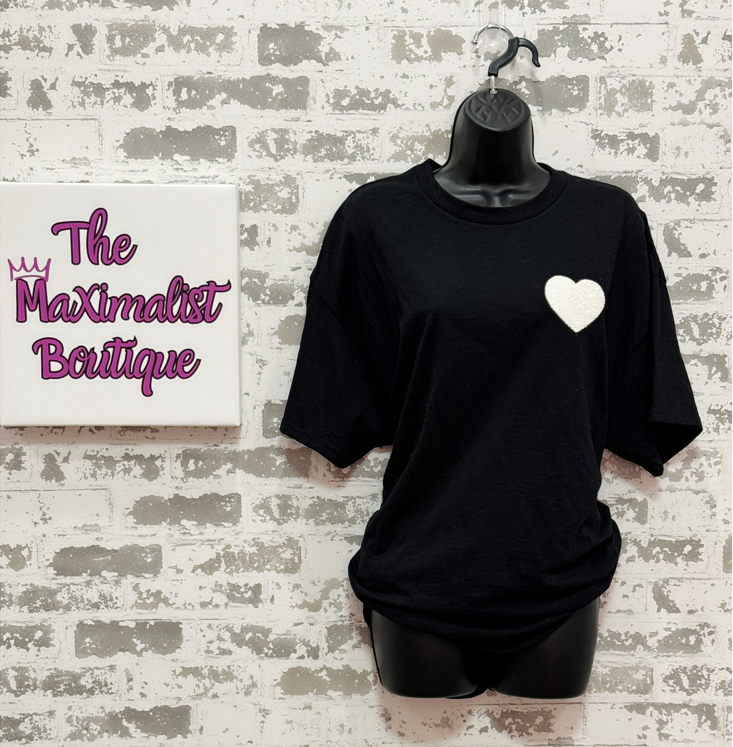 Black t-shirt with a white heart design on a mannequin against a textured wall with 'The Maximalist Boutique' branding.