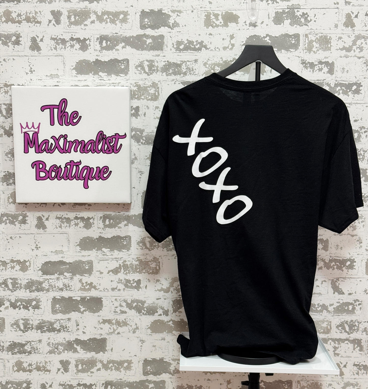 Black t-shirt with 'xoxo' on a hanger in front of 'The Maximalist Boutique' sign.