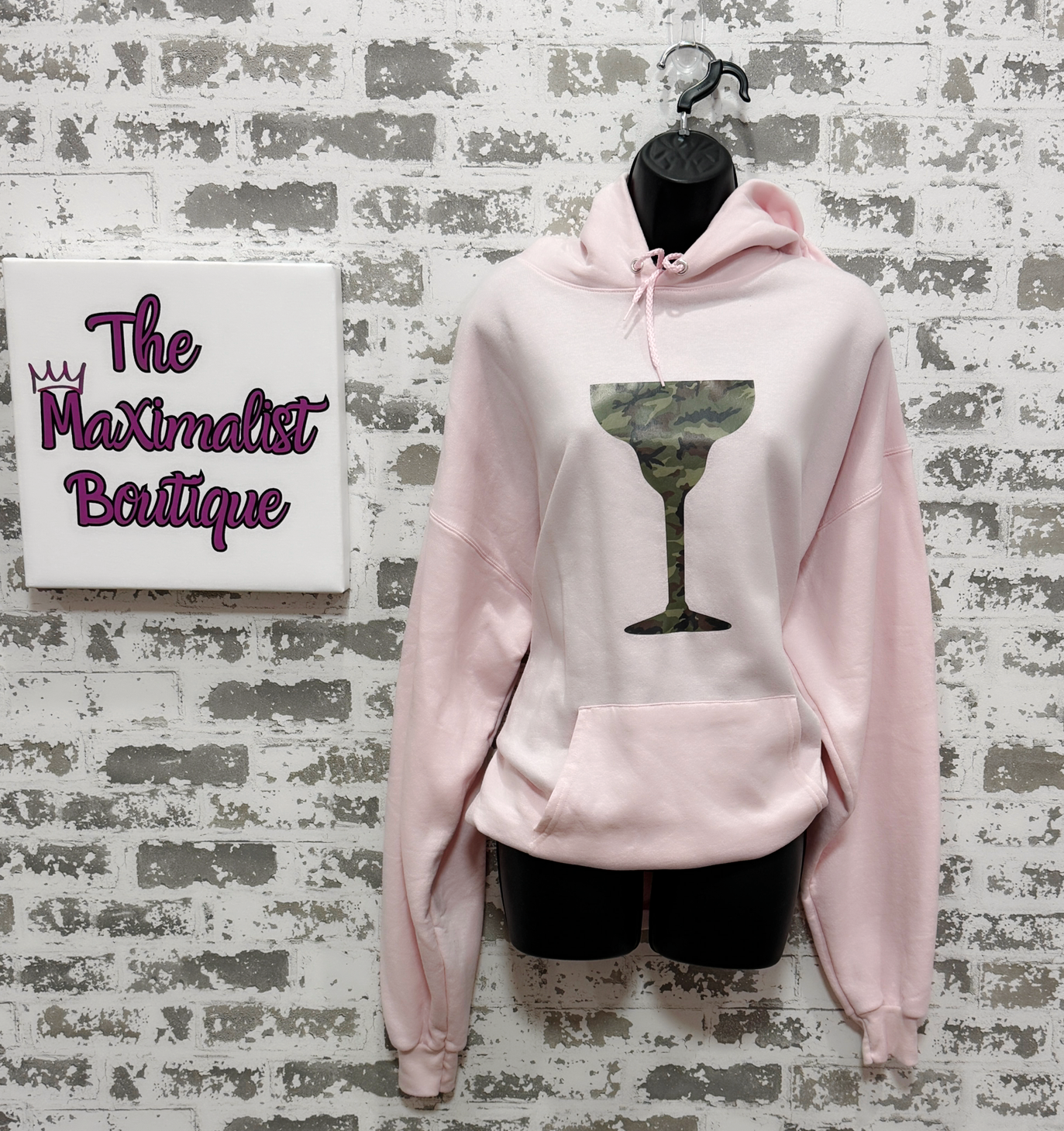 Pink hoodie with a graphic design on a textured wall with 'The Maximalist Boutique' sign.