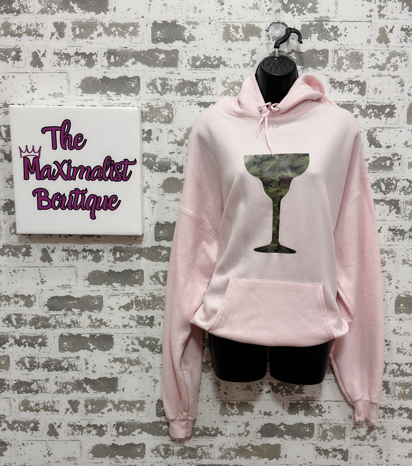 Pink hoodie with a camouflage cocktail graphic on a textured wall with 'The Maximalist Boutique' sign.