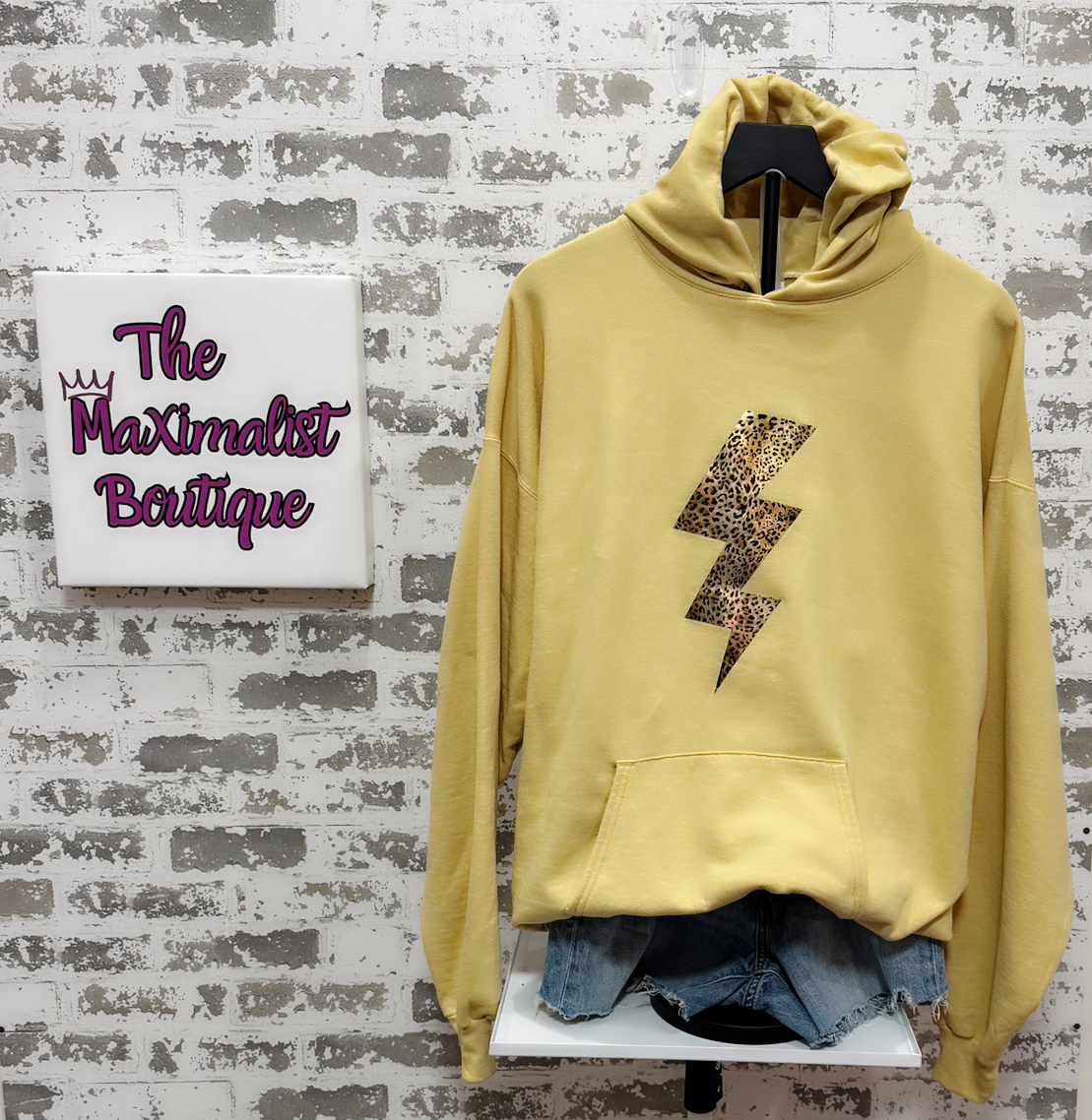 Yellow hoodie with a leopard print lightning bolt design on a stand against a textured wall with 'The Maximalist Boutique' sign.