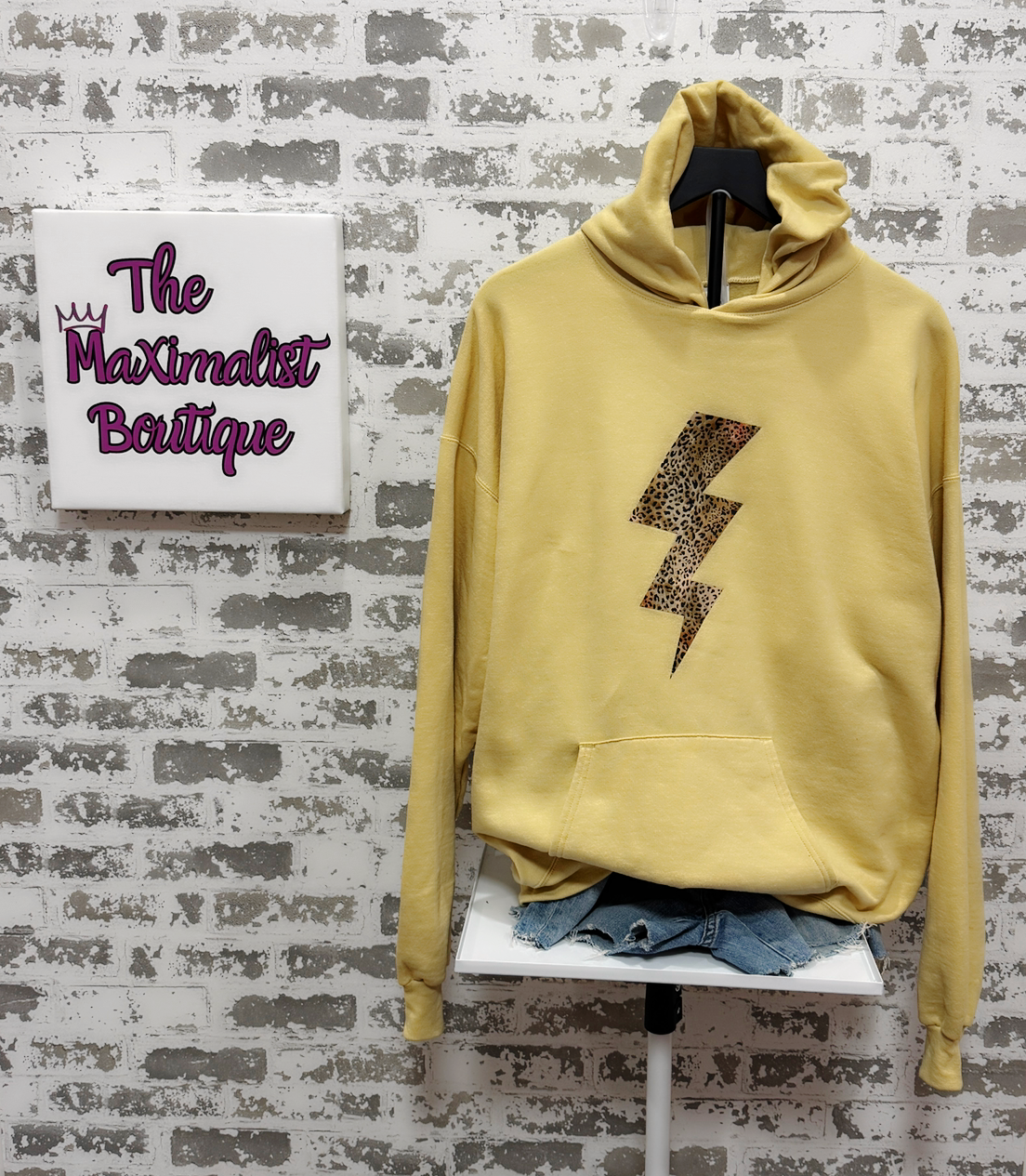 Yellow hoodie with a lightning bolt design on a chair against a textured wall with 'The Maximalist Boutique' sign.