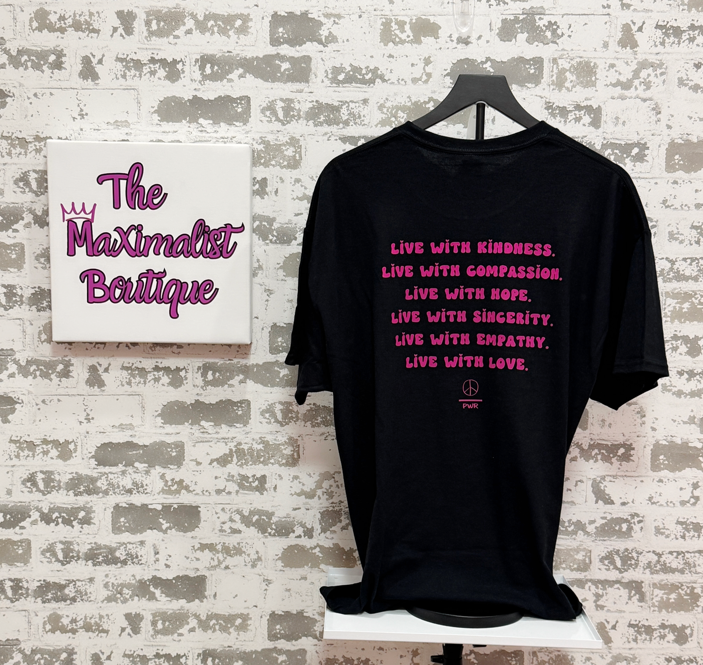 Black t-shirt with pink text on a hanger against a textured wall with 'The Maximalist Boutique' sign.