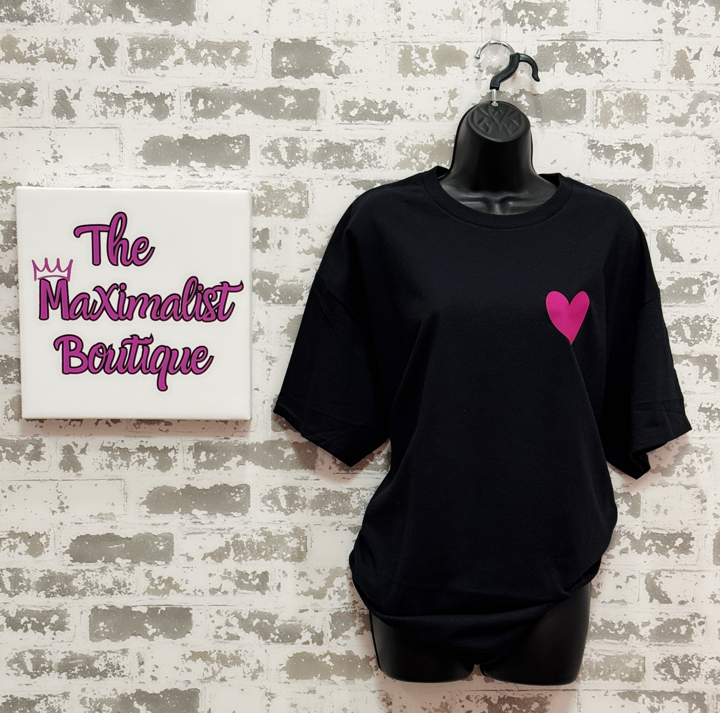 Black t-shirt with a pink heart and black shorts on a hanger against a textured wall with 'The Maximalist Boutique' sign.