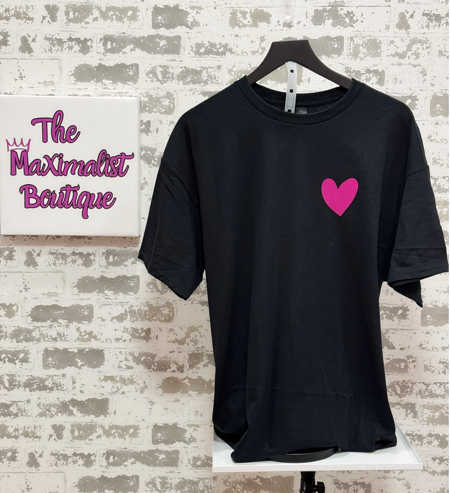 Black t-shirt with a pink heart on a hanger against a textured wall with 'The Maximalist Boutique' branding.