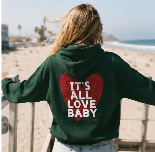 A woman standing on a beach railing, seen from behind, wearing a dark green hoodie with a large red glitter heart on the back featuring the white text 'IT'S ALL LOVE BABY'.