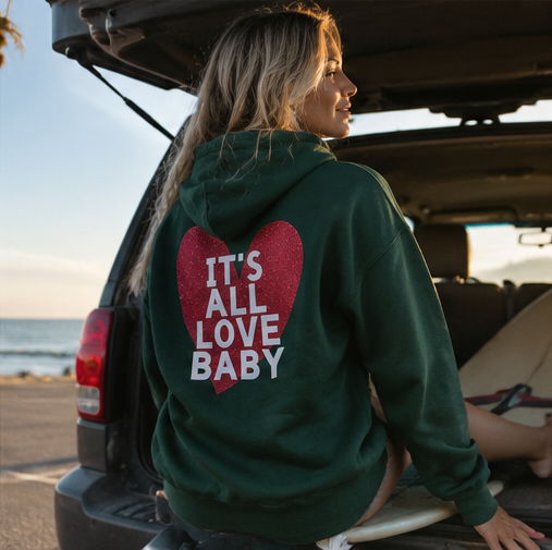 A woman sitting in the trunk of a vehicle by the beach wears a dark green hoodie with a red glitter heart and the text 'IT'S ALL LOVE BABY' on the back.