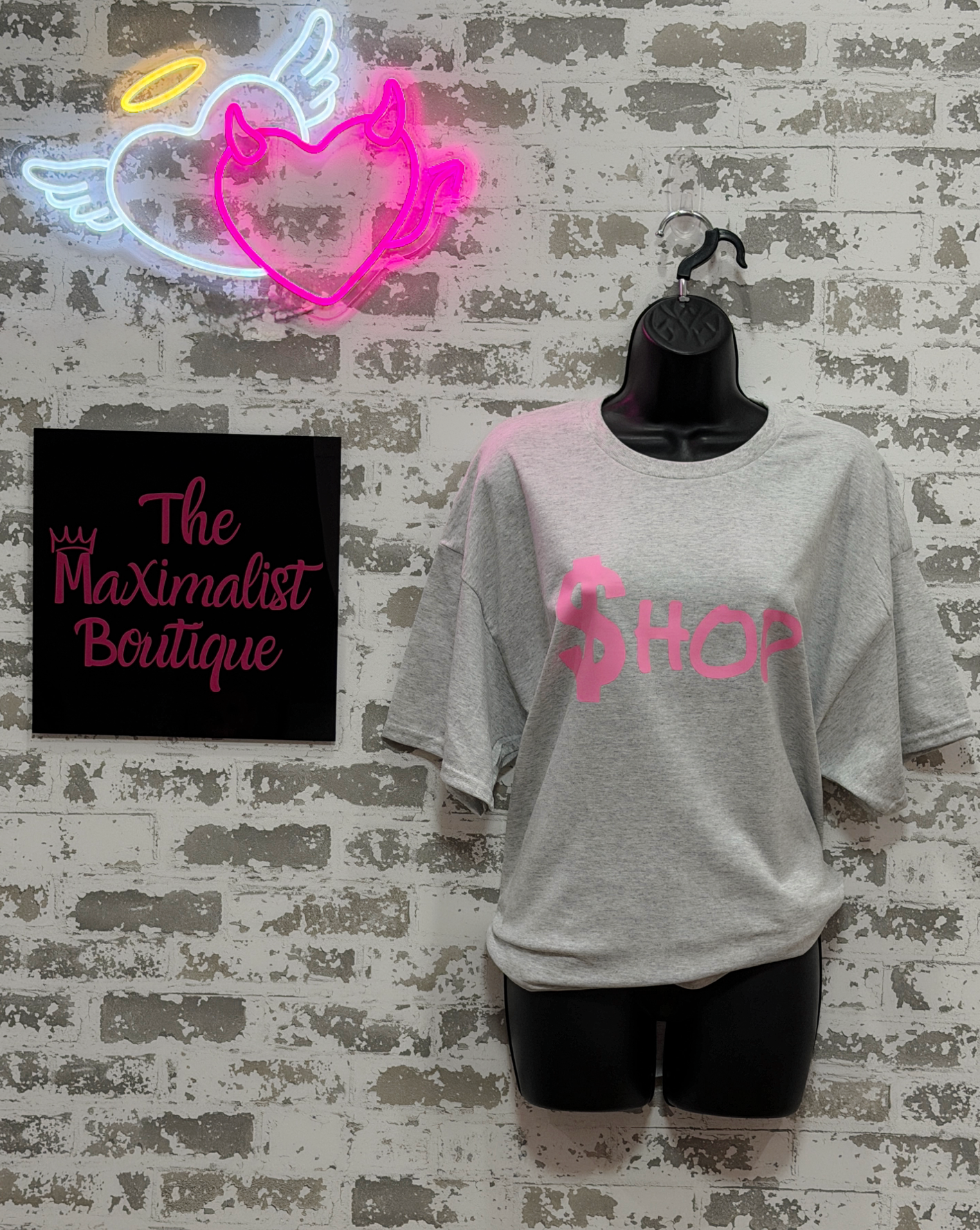 Gray t-shirt with pink text on a mannequin against a textured wall with neon sign and signboard.