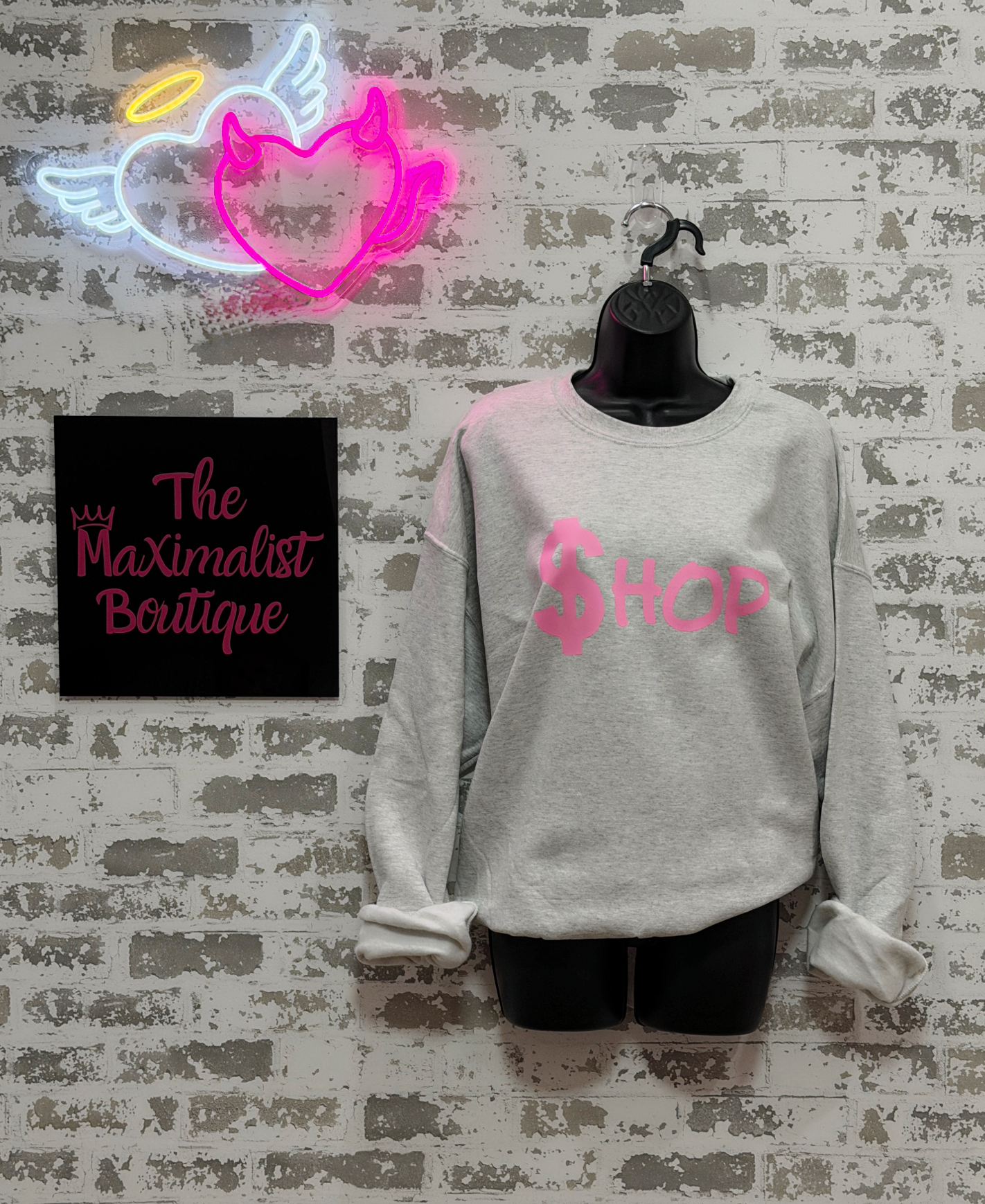 Gray sweatshirt with pink text on a mannequin against a textured wall with neon sign and 'The Maximalist Boutique' sign.