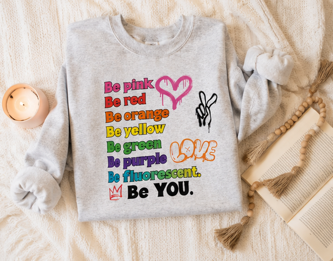 A light gray sweatshirt laid flat on a cream knit blanket, decorated with a colorful text graphic reading 'Be pink, red, orange, yellow, green, purple, fluorescent. Be YOU.
