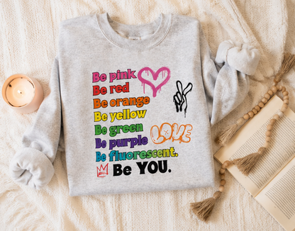 A light gray sweatshirt laid flat on a cream knit blanket, decorated with a colorful text graphic reading 'Be pink, red, orange, yellow, green, purple, fluorescent. Be YOU.