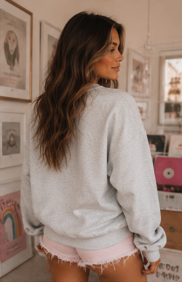 A woman with long brown hair is shown from behind wearing a light gray heather crewneck sweatshirt and light pink frayed denim shorts in a decorated room with framed art.