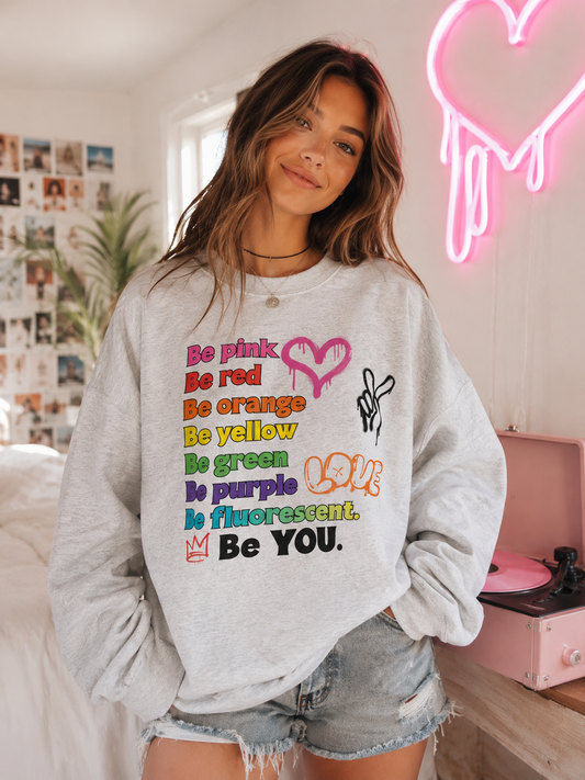 A woman wearing a light gray heather sweatshirt with a colorful graphic text print that reads 'Be pink, Be red, Be orange, Be yellow, Be green, Be purple, Be fluorescent. Be YOU.