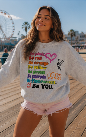 A woman wearing a light gray sweatshirt with colorful retro text that reads 'Be pink, Be red, Be orange, Be yellow, Be green, Be purple, Be fluorescent. Be YOU.