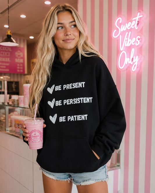 A woman wearing a black hoodie with the white printed text 'BE PRESENT', 'BE PERSISTENT', and 'BE PATIENT' stands in a pink-and-white striped cafe holding a pink drink.
