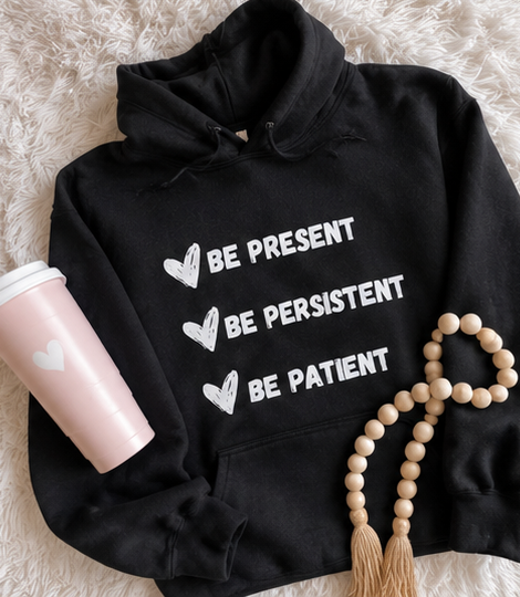A black pullover hoodie laid flat on a white faux-fur surface, printed with three white heart-and-text phrases reading 'BE PRESENT,' 'BE PERSISTENT,' and 'BE PATIENT.' A pink tumbler with a white heart, a beige wooden bead necklace, and a neutral blanket are styled around it.