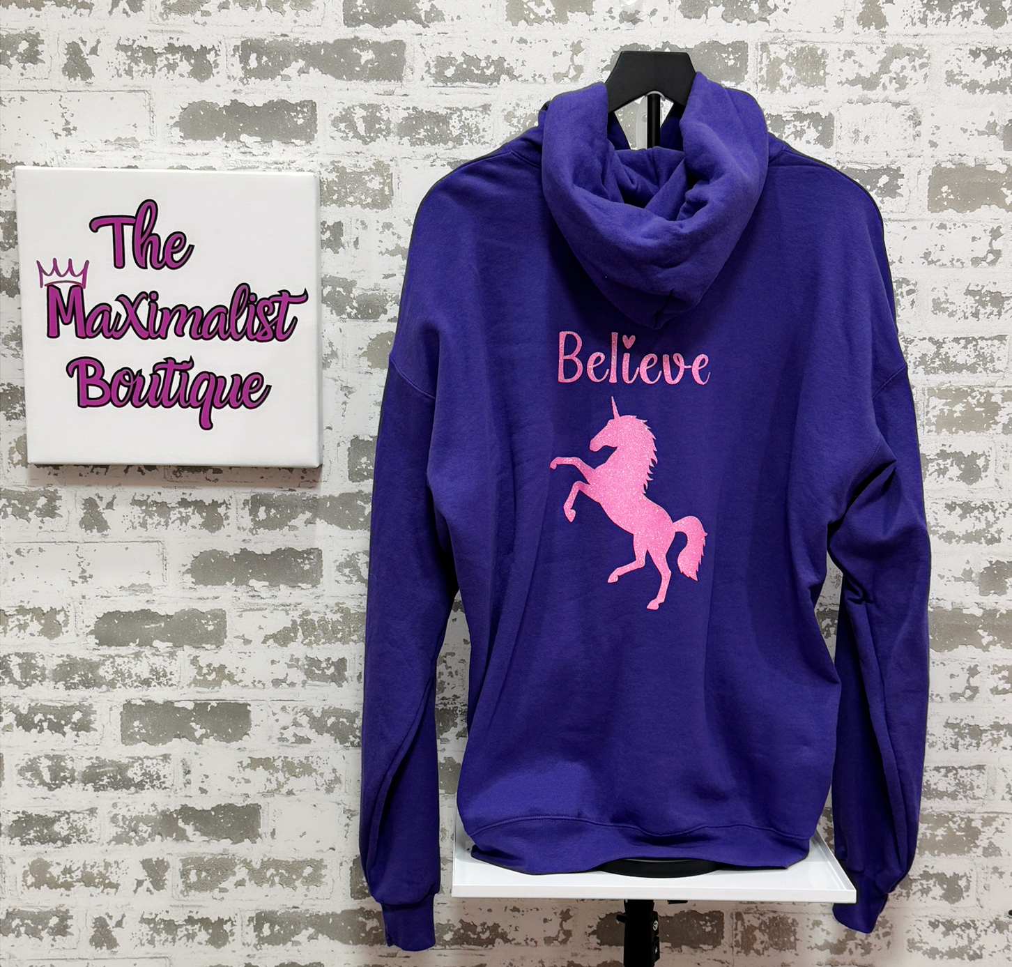 Purple hoodie with pink unicorn and text on a white brick wall background