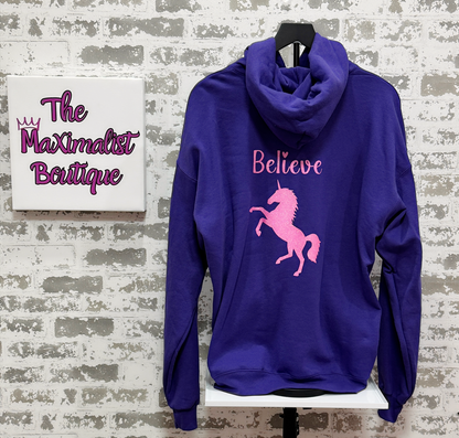 Purple hoodie with pink unicorn and text on a white brick wall background