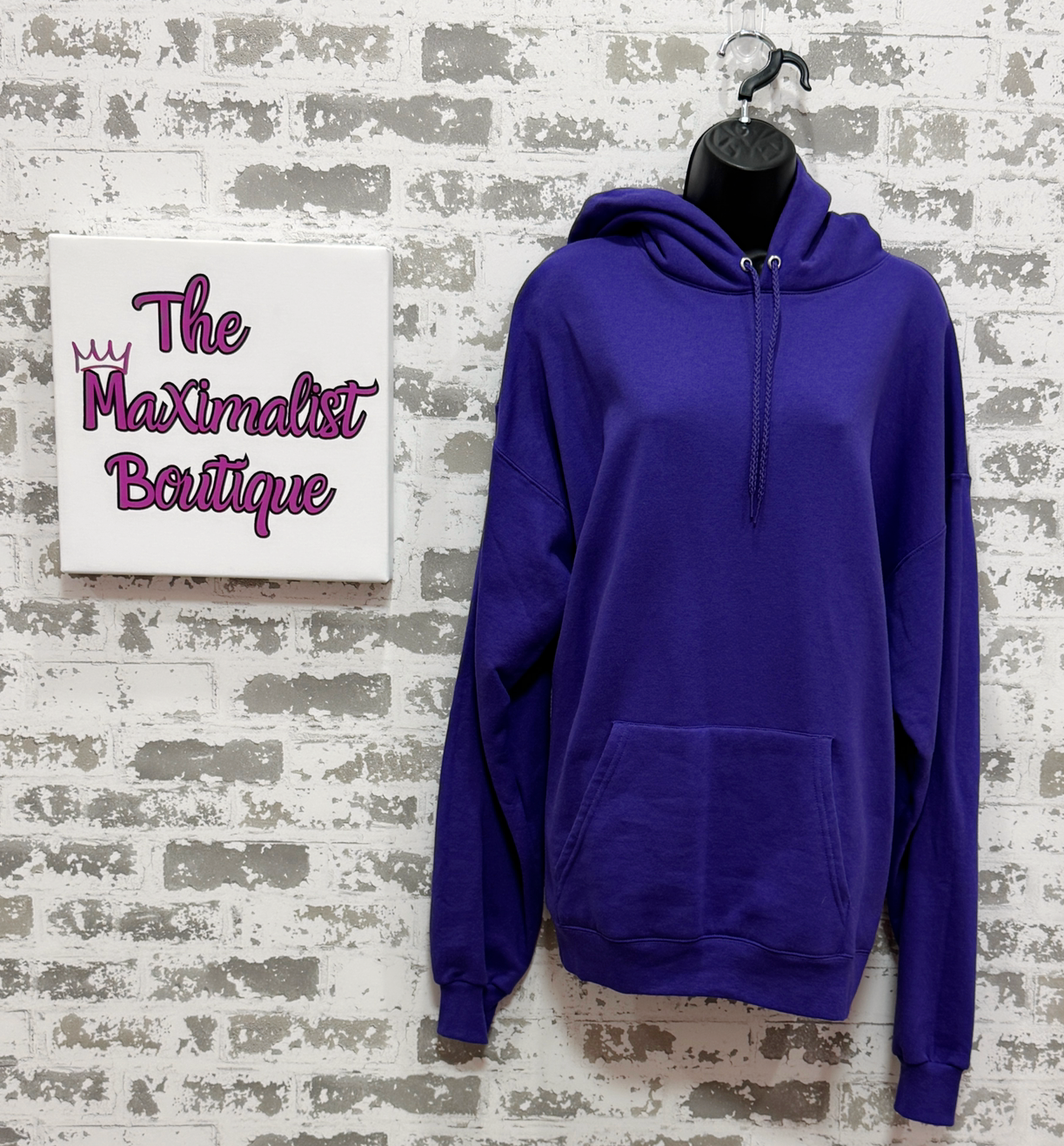 Purple hoodie on a mannequin against a textured wall with 'The Maximalist Boutique' sign.
