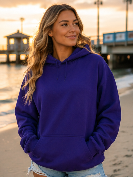 A woman wearing a solid purple pullover hoodie stands on a beach at sunset, with a pier and water blurred in the background.