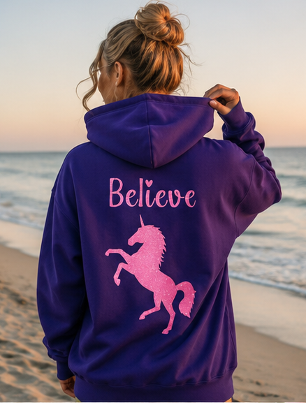 A woman standing on a beach at sunset, viewed from behind, wearing a purple hoodie with a pink unicorn graphic and the word 'Believe' printed on the back.