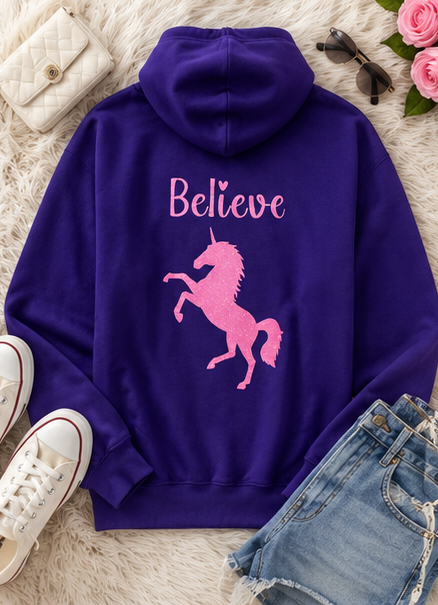 A purple hoodie laid flat on a white faux-fur surface, featuring a pink unicorn graphic and the word 'Believe' in pink across the back. The hoodie is styled with white sneakers, blue denim shorts, sunglasses, and a quilted cream handbag.
