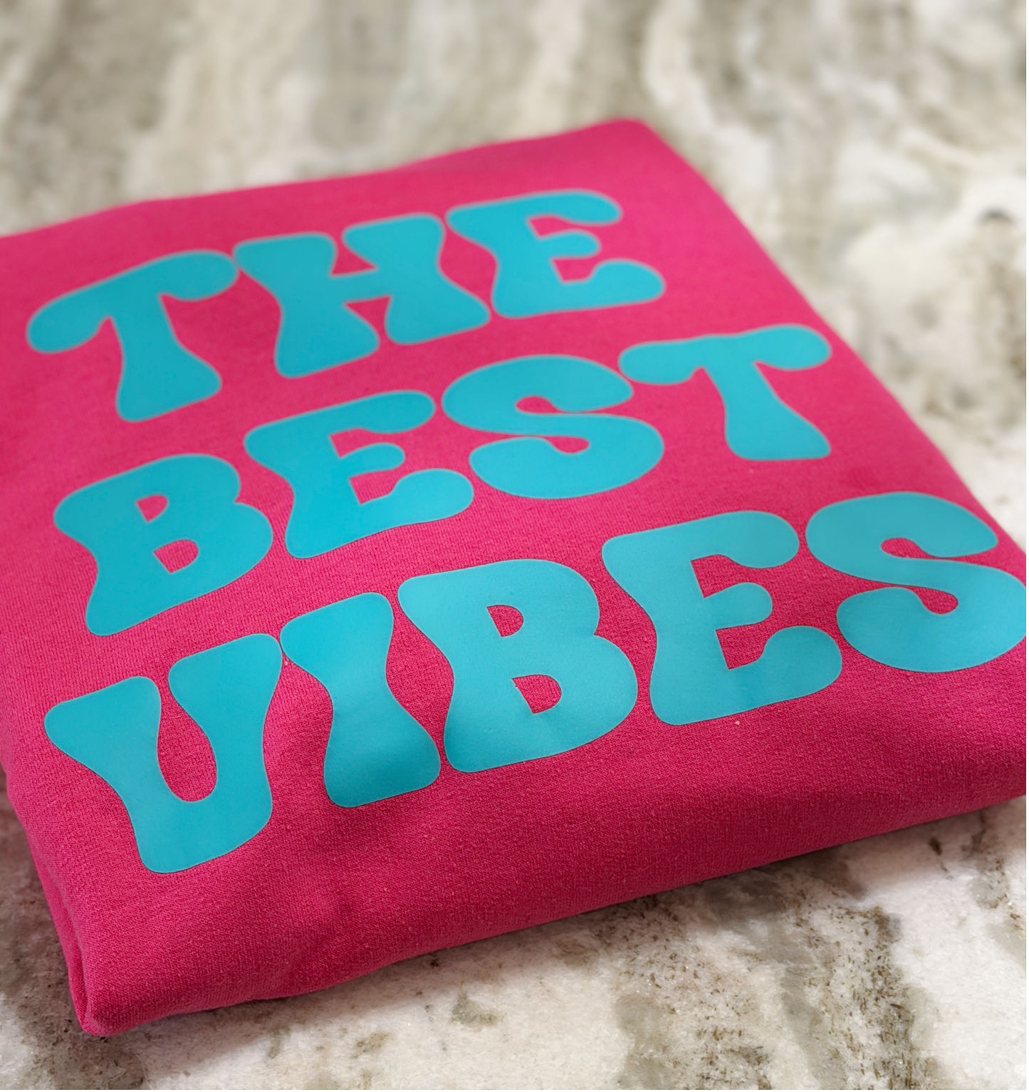 Pink shirt with 'THE BEST VIBES' text in blue on a textured surface
