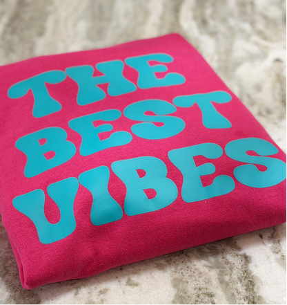 Pink shirt with 'THE BEST VIBES' text in blue on a textured surface