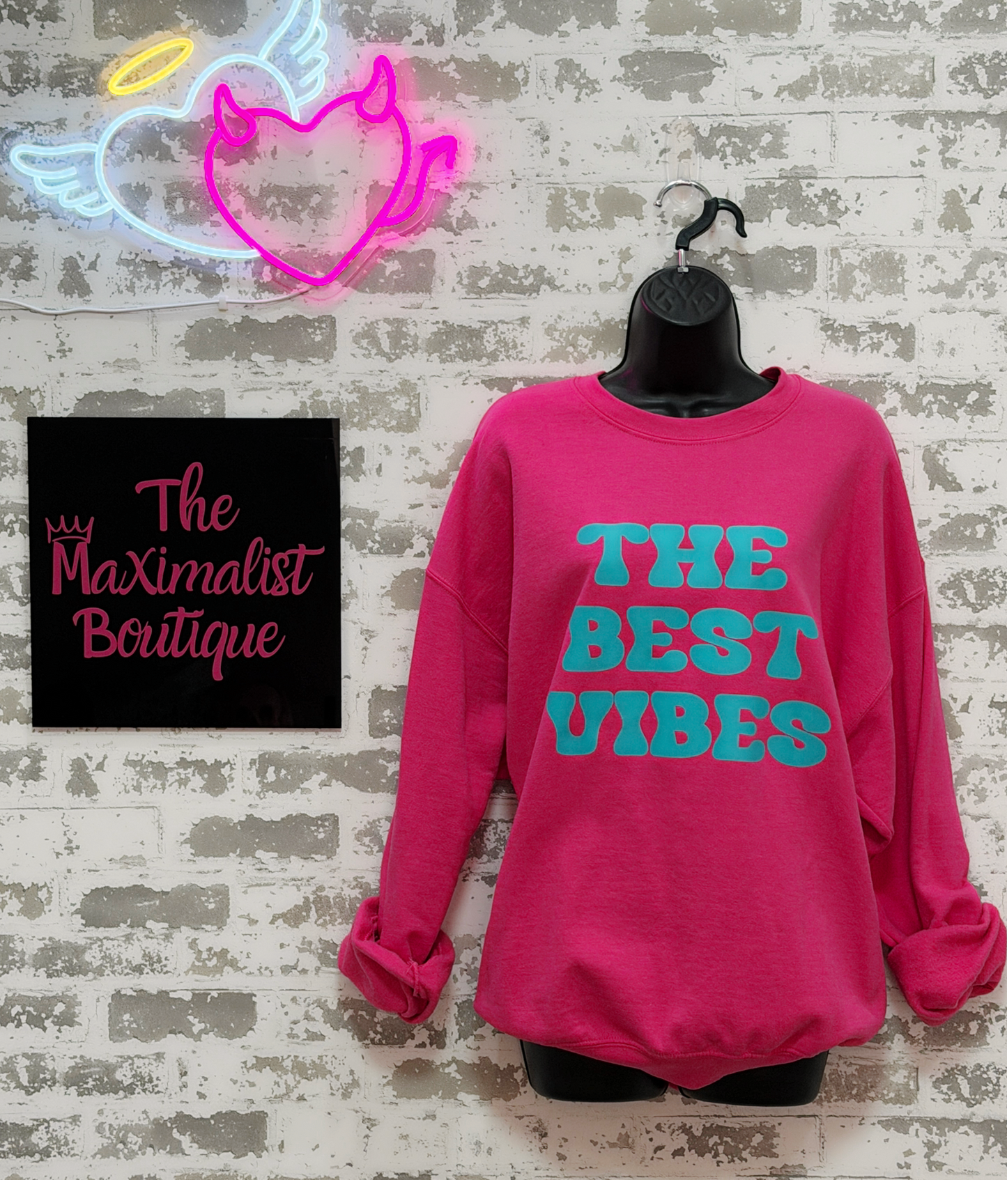 Pink sweatshirt with 'The Best Vibes' text on a textured wall background with 'The Maximalist Boutique' sign.