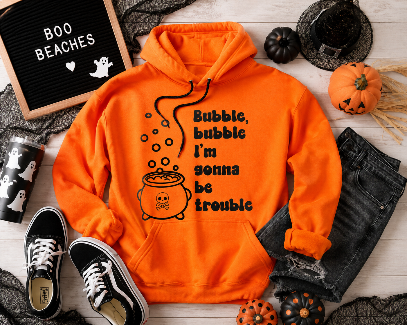 Bright orange pullover hoodie with a black graphic of a cauldron and the text 'Bubble, bubble I'm gonna be trouble,' styled in a flat lay with black jeans, black sneakers, small pumpkins, a witch hat, lace, and a letter board that says 'BOO BEACHES'.